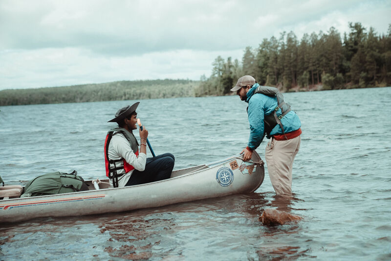 A man in a canoe is talking to another man standing in the water. The man in the canoe is wearing a life vest and holding a paddle. The man in the water is wearing a hat and a jacket. They are in a lake or river, and there are trees in the background.